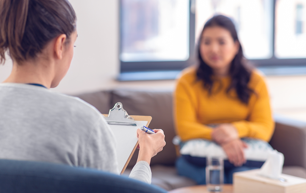 Das Foto zeigt eine Szene in einer psychotherapeutischen Praxis. Rechts ist eine Patientin zu sehen, die auf einer brauen Couch sitzt. Links im Bild befindet sich die Psychotherapeutin, die auf einem Stuhl sitzt und das Gespräch handschriftlich auf einem Protokollbogen dokumentiert.